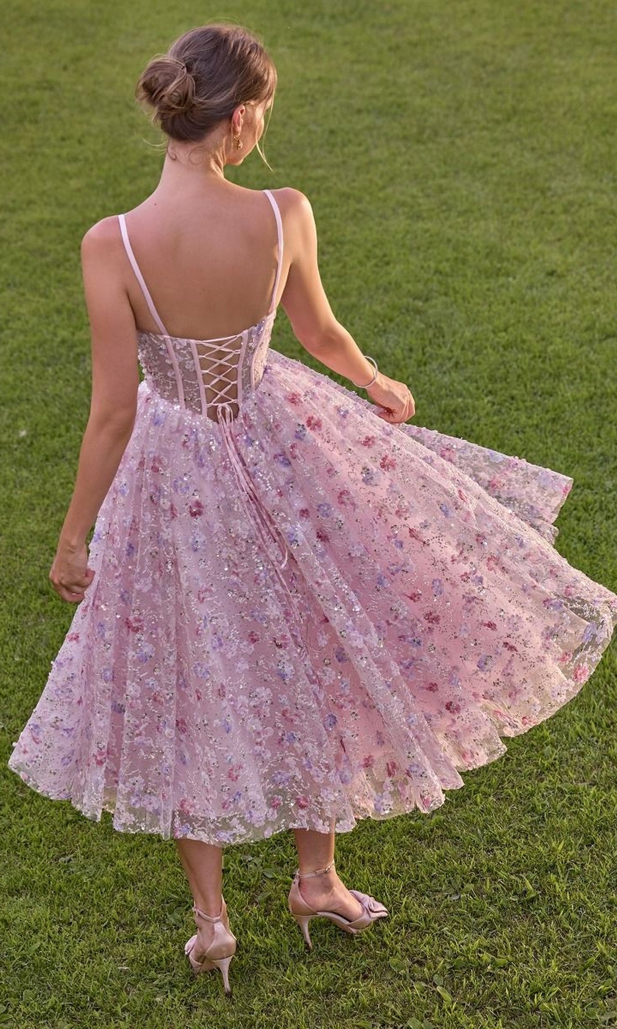 A woman stands on grass wearing the Andrea & Leo A1523 midi prom dress, a light pink floral lace design featuring a sheer corset bodice with a lace-up back and delicate spaghetti straps, paired with matching heels.